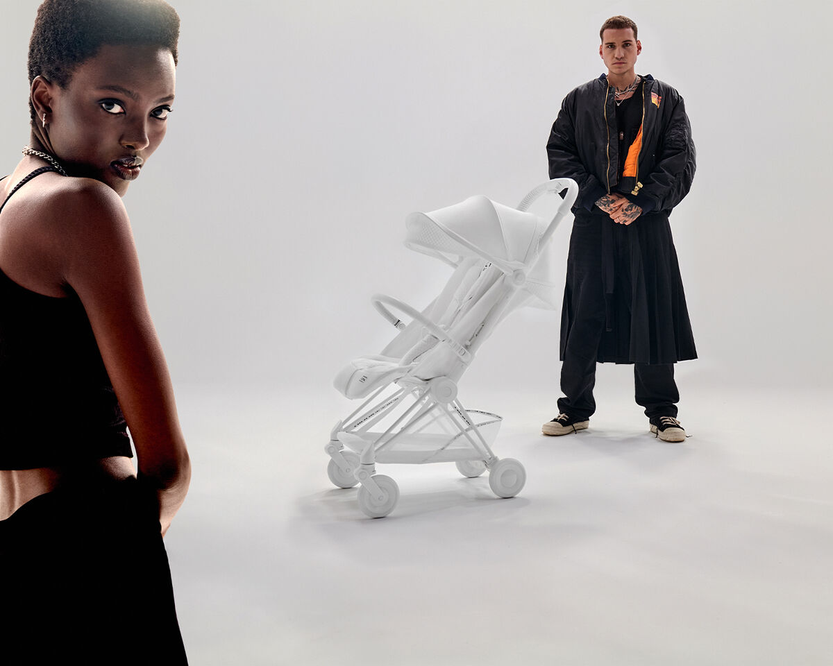 A woman in a black top stands in the foreground on the left, while a man in a long black coat and trainers stands next to a white pushchair in the background.
