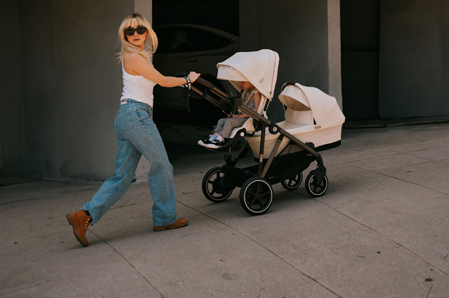 A woman in jeans and a white tank top pushes a stroller with two children on a pavement.