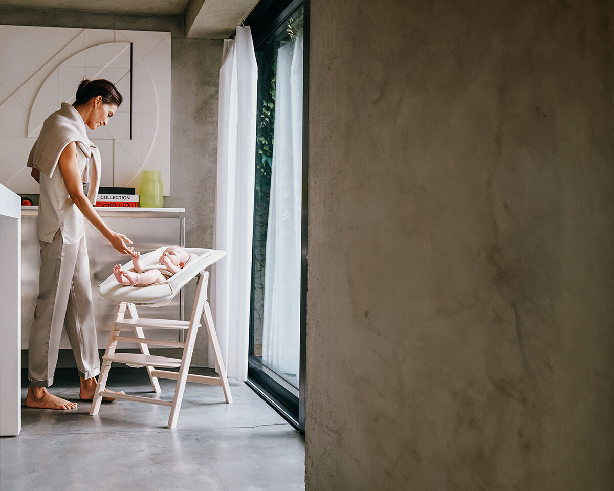 A woman stands in a modern kitchen, smiling at a baby lying in a high chair.