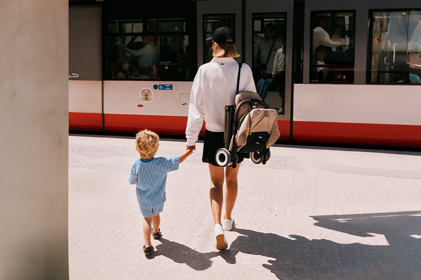 Madre e bambino che camminano mano nella mano davanti a un tram. La madre porta un passeggino piegato.