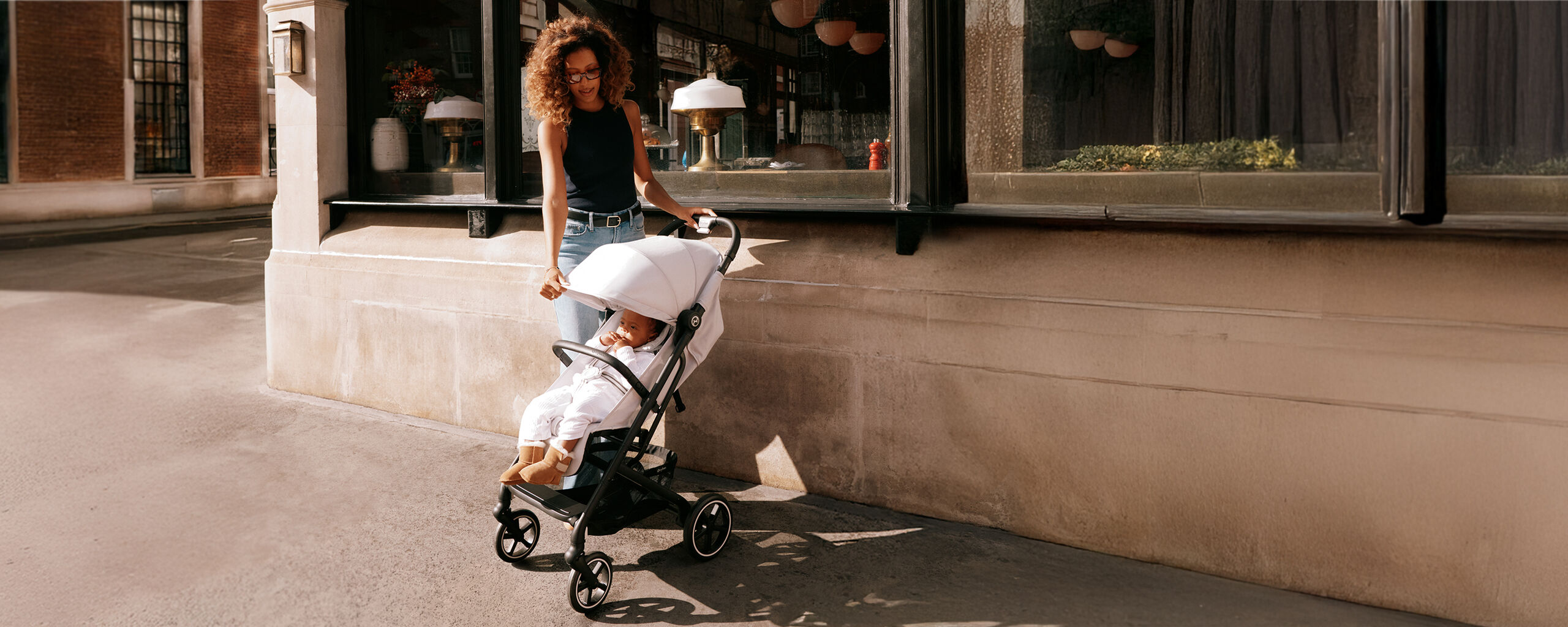 A woman with curly hair is pushing the Beezy lightweight compact stroller, with a baby sitting inside.
