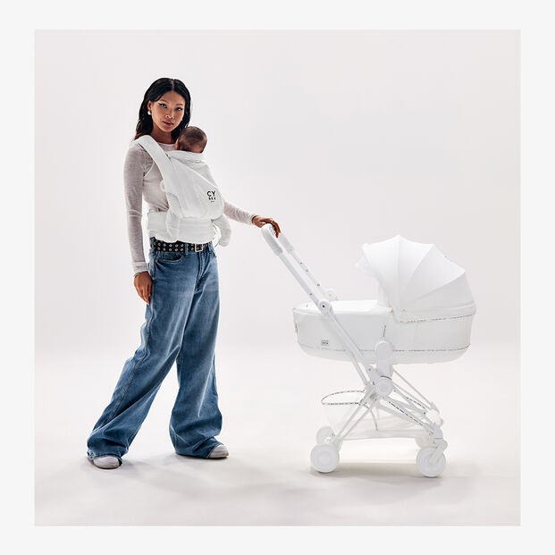 Una donna con una mano su un passeggino Urban Mobility, mentre tiene il suo bambino in un marsupio Urban Mobility.