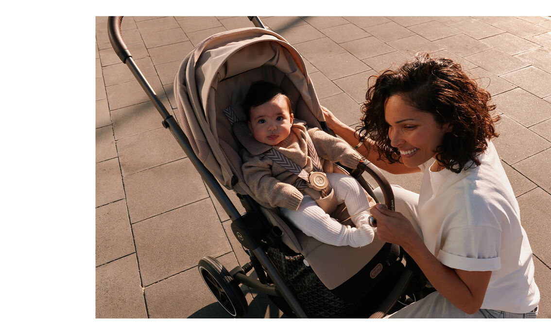 A baby sitting in a Balios S Lux stroller with seat pack and looking into the camera while the mother is kneeling in front with a grin.