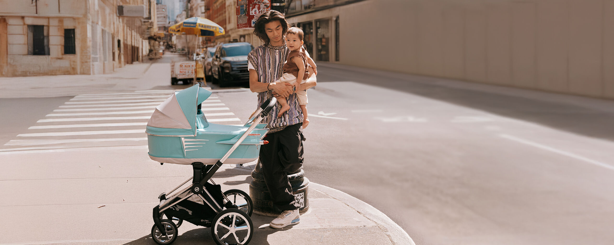 A young father is holding a child in his arms while standing next to a modern stroller on a city street.
