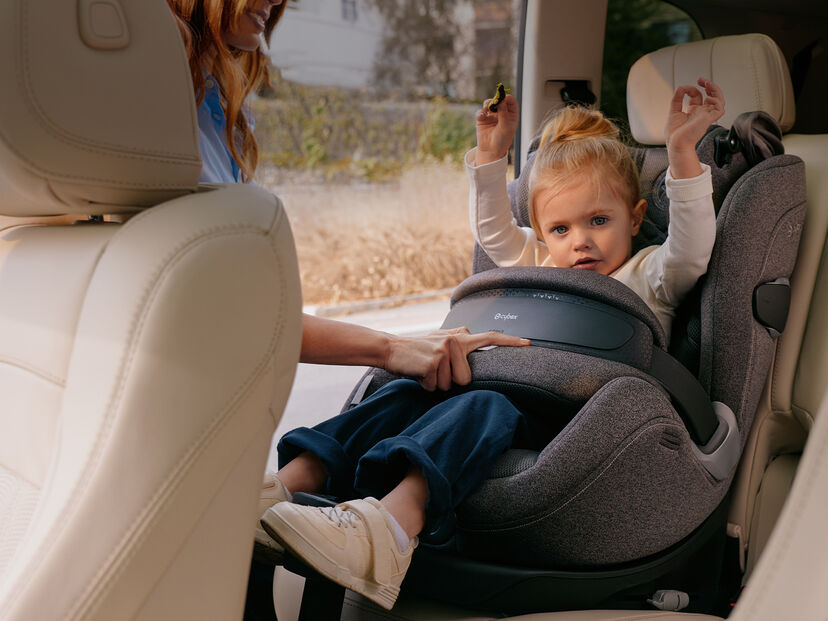A small child sits in a Cybex car seat in a vehicle while a woman adjusts the safety belt.