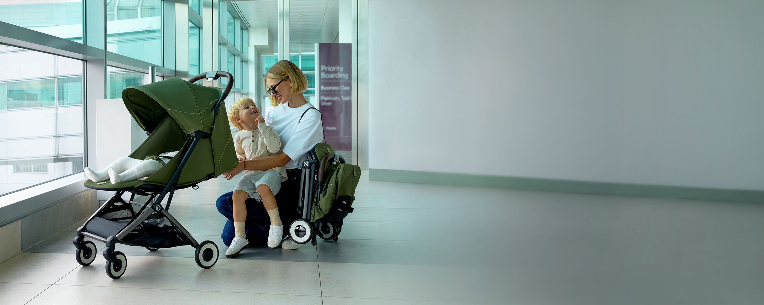 A compact travel stroller named Orfeo in a modern airport, a mother sits on the floor with her child while the stroller is beside them.