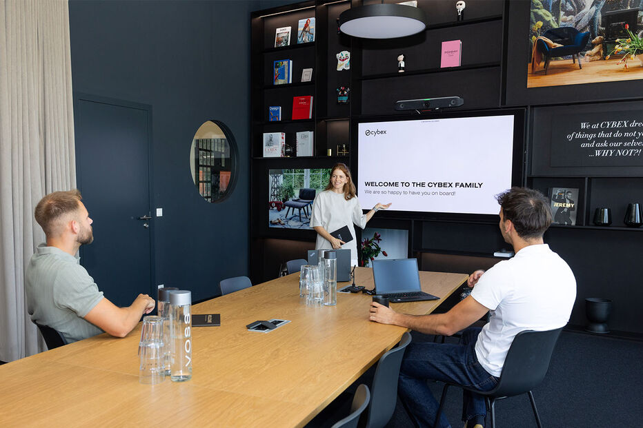 Three colleagues in a meeting room &mdash; one woman presenting at a screen, two men seated at the table listening.