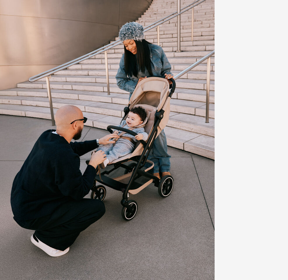 Une m&egrave;re debout derri&egrave;re sa poussette compacte Beezy, tenant le guidon. Son enfant est &agrave; l&rsquo;int&eacute;rieur, le p&egrave;re est agenouill&eacute; devant lui et chatouille le b&eacute;b&eacute;.