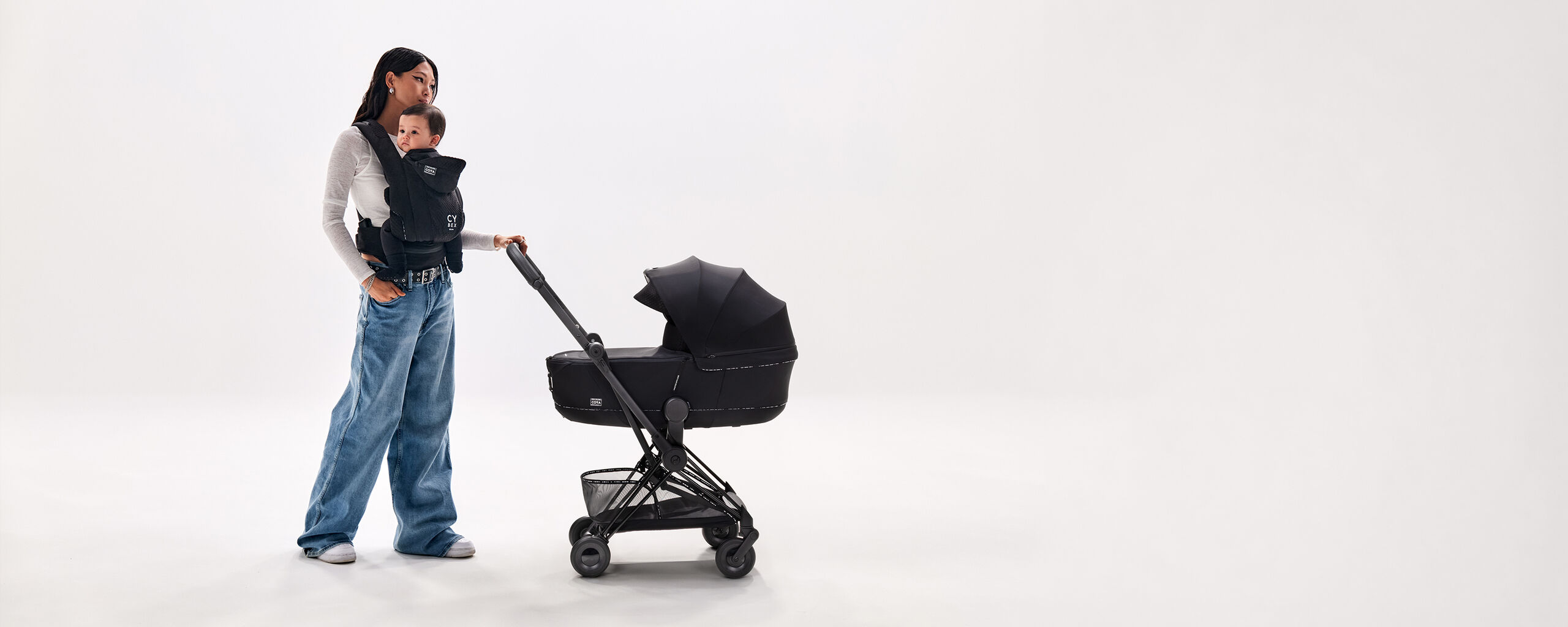 Una donna con una mano sul manubrio del suo Urban Mobility Coya nero mentre tiene il suo bambino in un marsupio Urban Mobility nero.