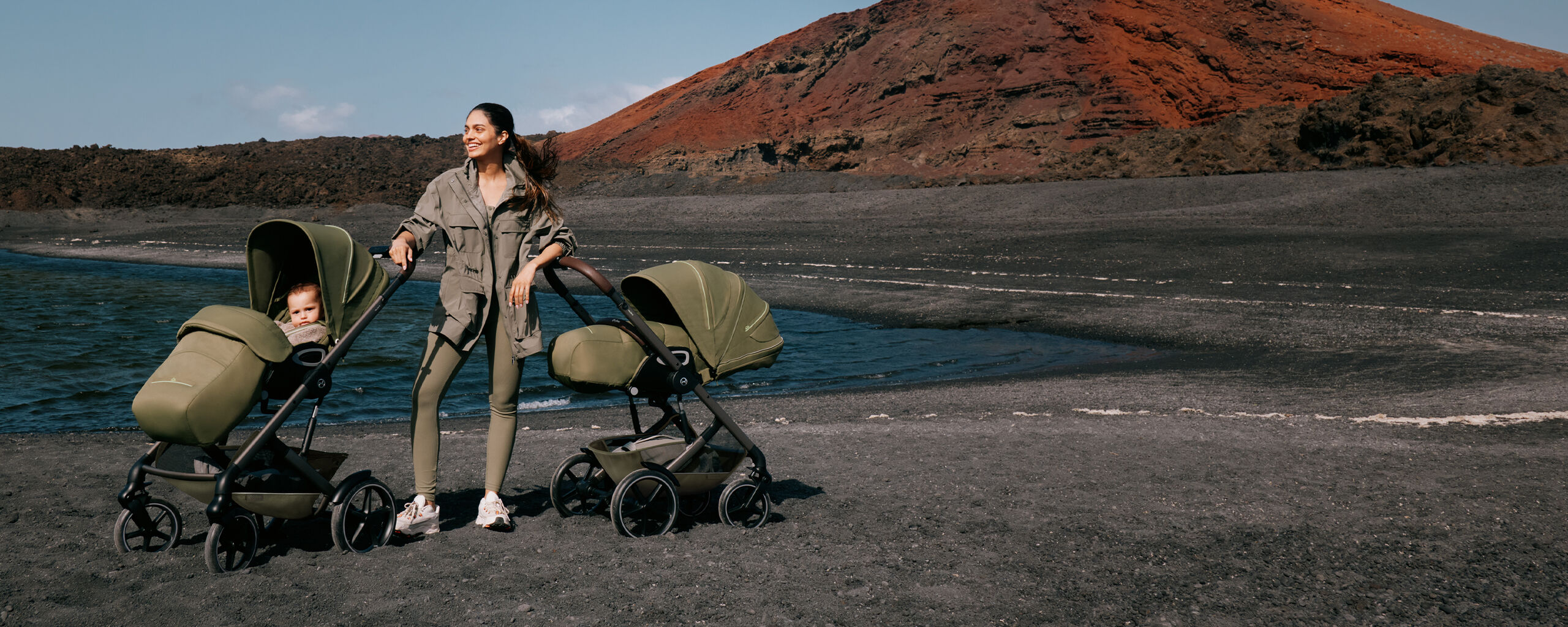 Eine Frau steht in schwarzem Sand am Strand mit zwei Talos S Lux Kinderwagen links und rechts neben ihr.