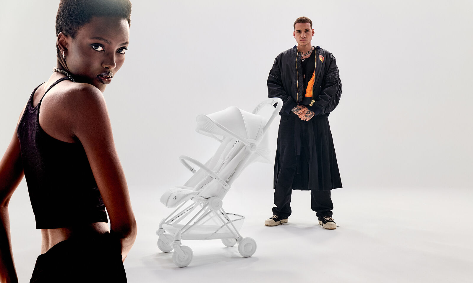 A woman in a black top stands in the foreground, while a man in black clothing stands next to a white stroller in the background.