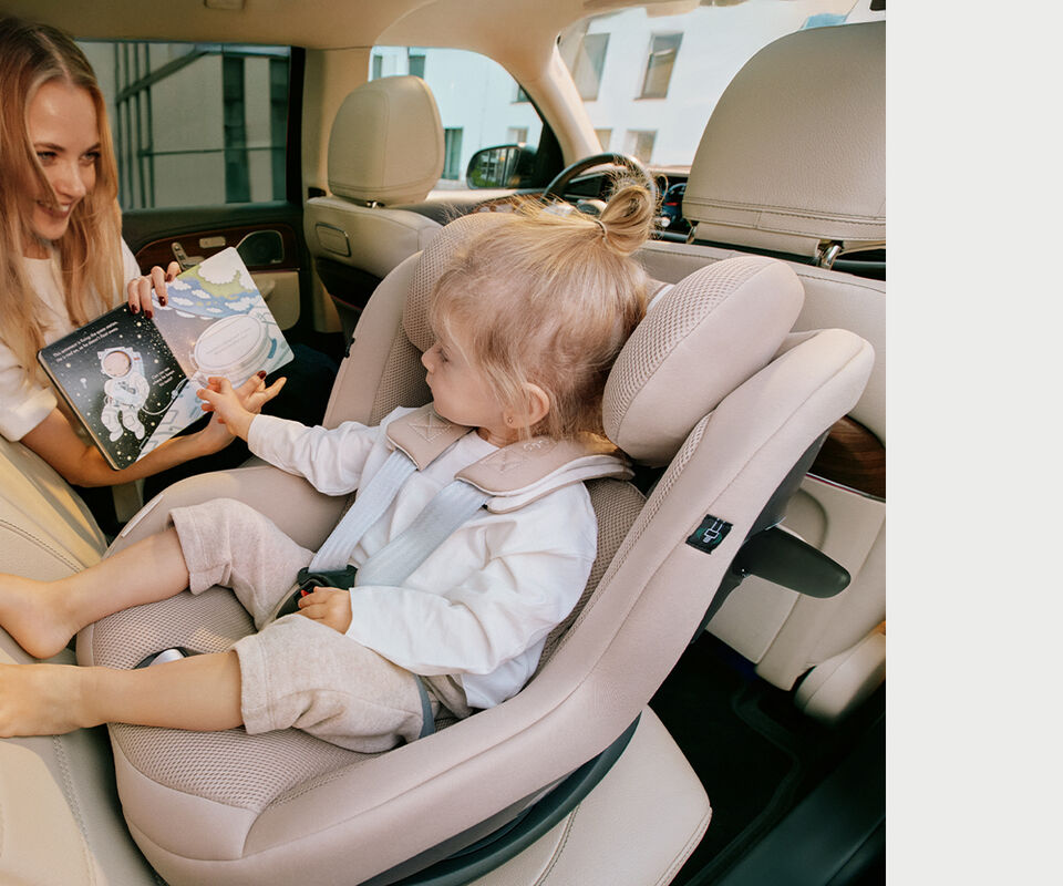 Woman sitting with her daughter in the back of a car. The daughter is sitting in a Sirona G3 car seat that is turned rear-facing while she looks at a picture book the mother is showing her.