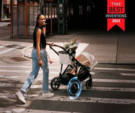 A woman with long hair wearing a black top and jeans walks across a crosswalk with a modern stroller. The stroller contains fresh groceries and features a futuristic design with glowing wheels.