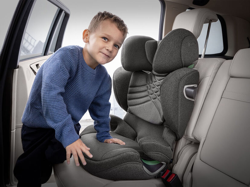 A child in a blue jumper stands next to a grey car seat in a vehicle.