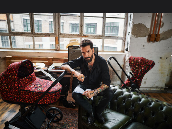 A man sitting in a loft-like room on a green leather sofa, surrounded by two CYBEX Rockstar strollers with a red pattern.
