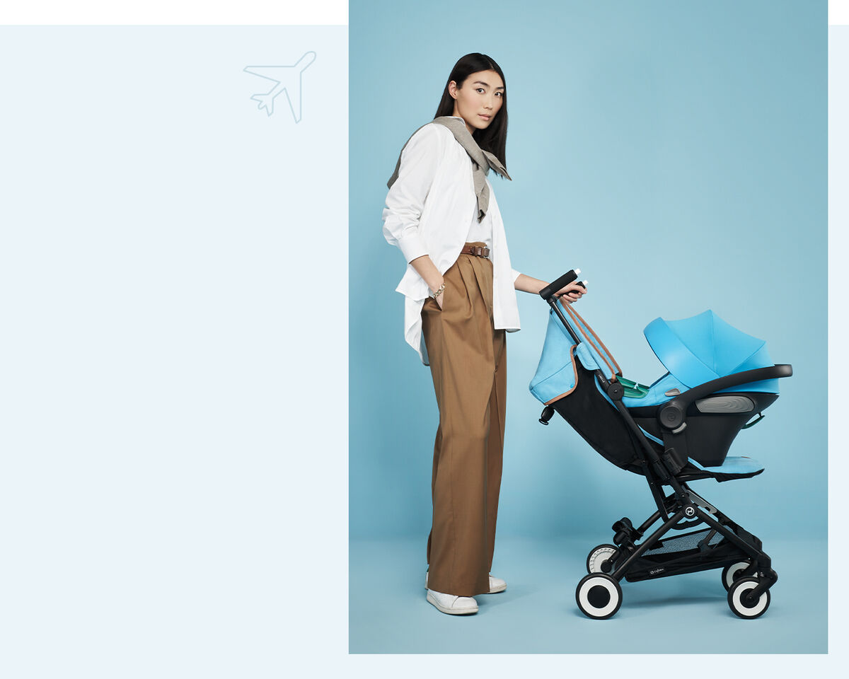 Woman in a white shirt and brown trousers stands next to a blue Libelle, the smallest fold stroller, against a blue background.
