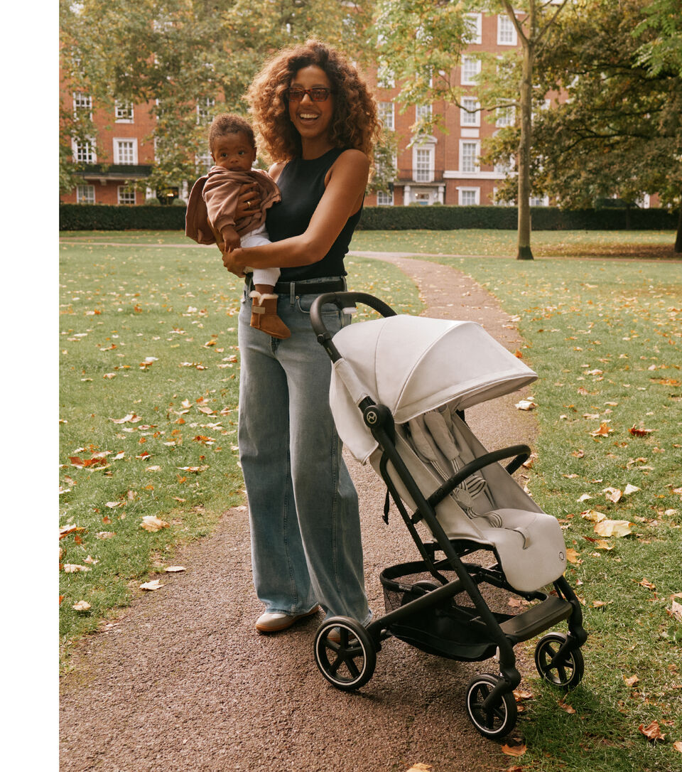 A woman with curly hair holds a baby in a park, next to her is the Beezy lightweight compact stroller.