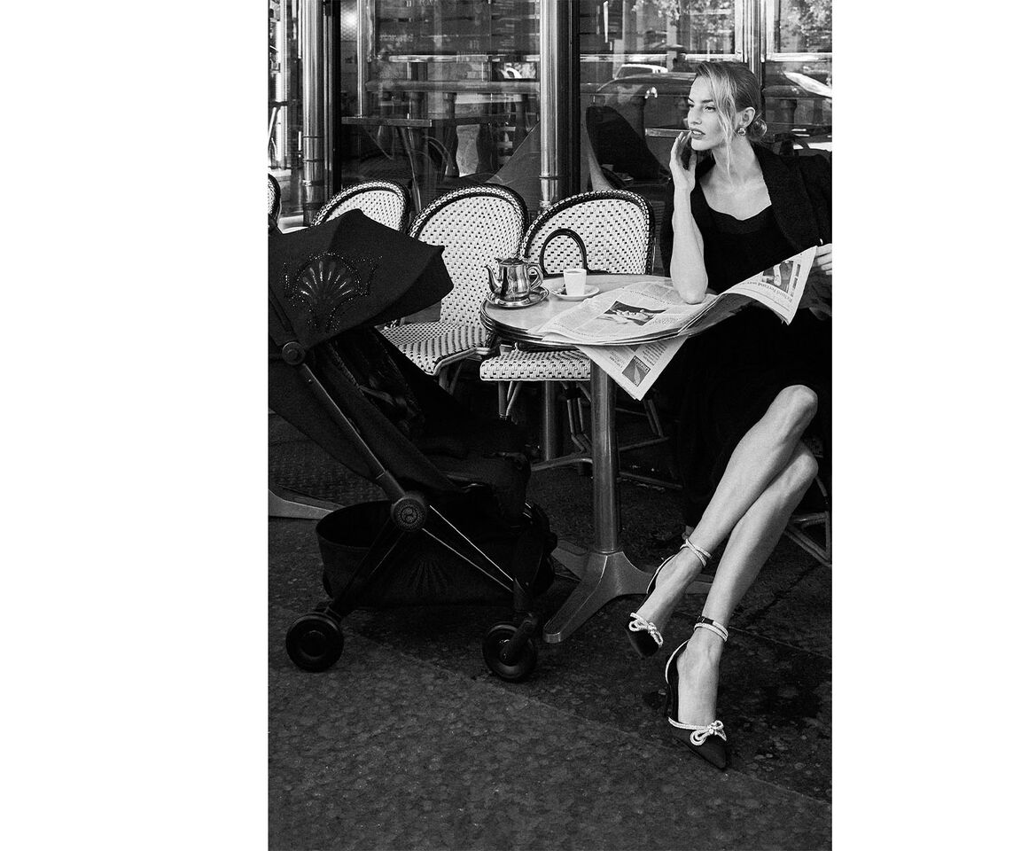 A woman sitting in a caf&eacute; reading a newspaper while talking on the phone. A stroller is next to her.