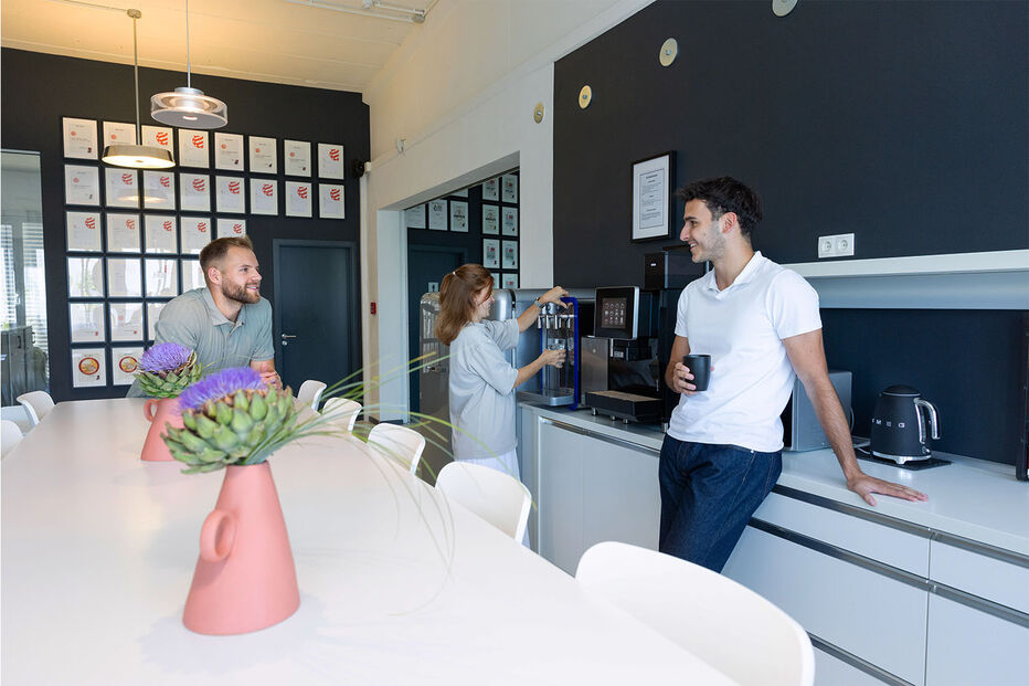 In an office kitchenette, two male colleagues chat while a third female colleague pours coffee.