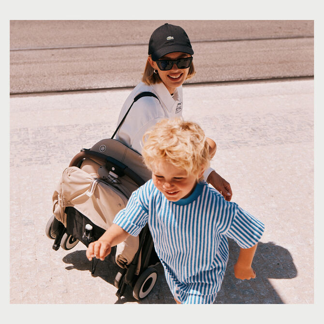 Eine Frau mit Baseball Cap und Sonnenbrille schaut ihrem Kind mit einem lachen beim Spielen zu. Die tr&auml;gt einen gefalteten CYBEX Buggy &uuml;ber die Schulter.