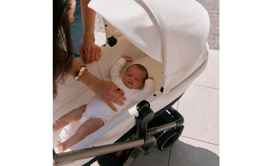 En nyf&ouml;dd ligger i en CYBEX-barnvagn med tyg i Seashell Beige. Mamman b&ouml;jer sig fram&aring;t medan hennes hand f&ouml;rsiktigt smeker bebisen.