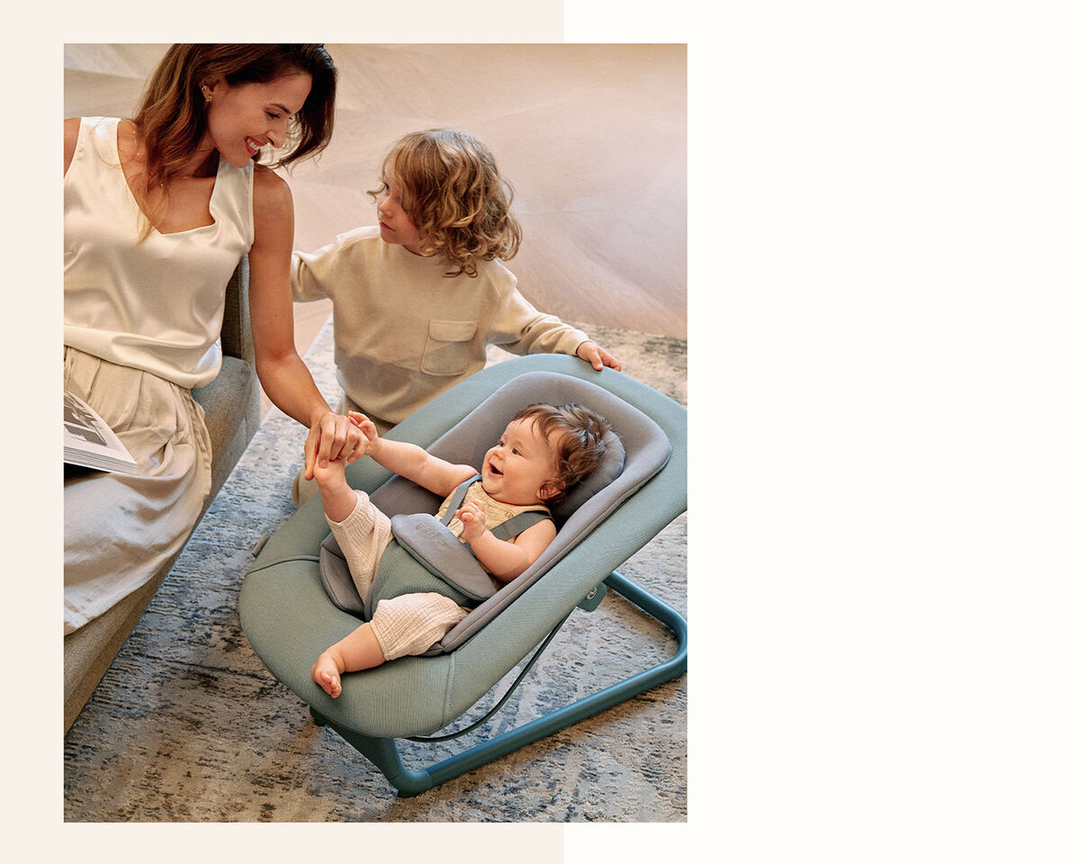 A mother sits on a sofa holding hands with her smiling child, while another child is seated in a modern blue baby chair.