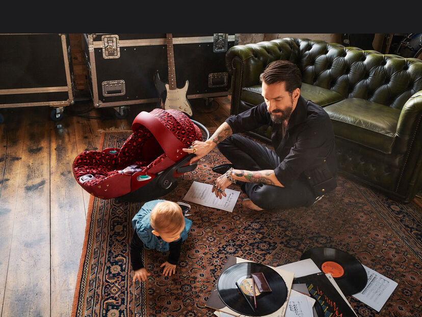 CYBEX Rockstar Babyschale in einem Wohnzimmer mit einem Mann und einem krabbelnden Baby auf einem Teppich.