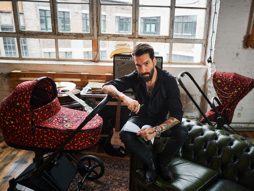 En sk&auml;ggig man med tatueringar sitter p&aring; en gr&ouml;n l&auml;dersoffa i ett rum med stora f&ouml;nster. Bredvid honom st&aring;r en barnvagn med r&ouml;tt leopardm&ouml;nster.
