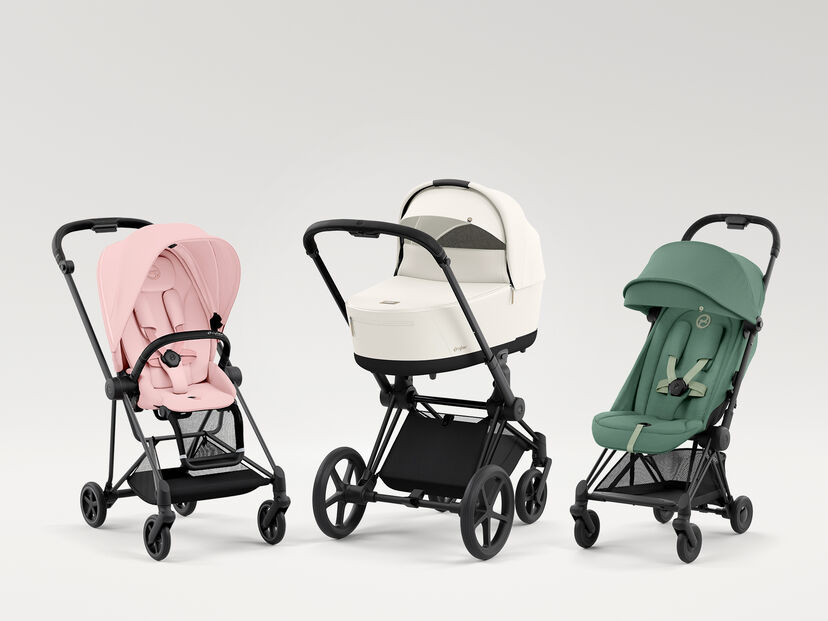 Three pushchairs in different colours: a pink pushchair on the left, a white pushchair with a carrycot in the middle, and a green pushchair on the right.