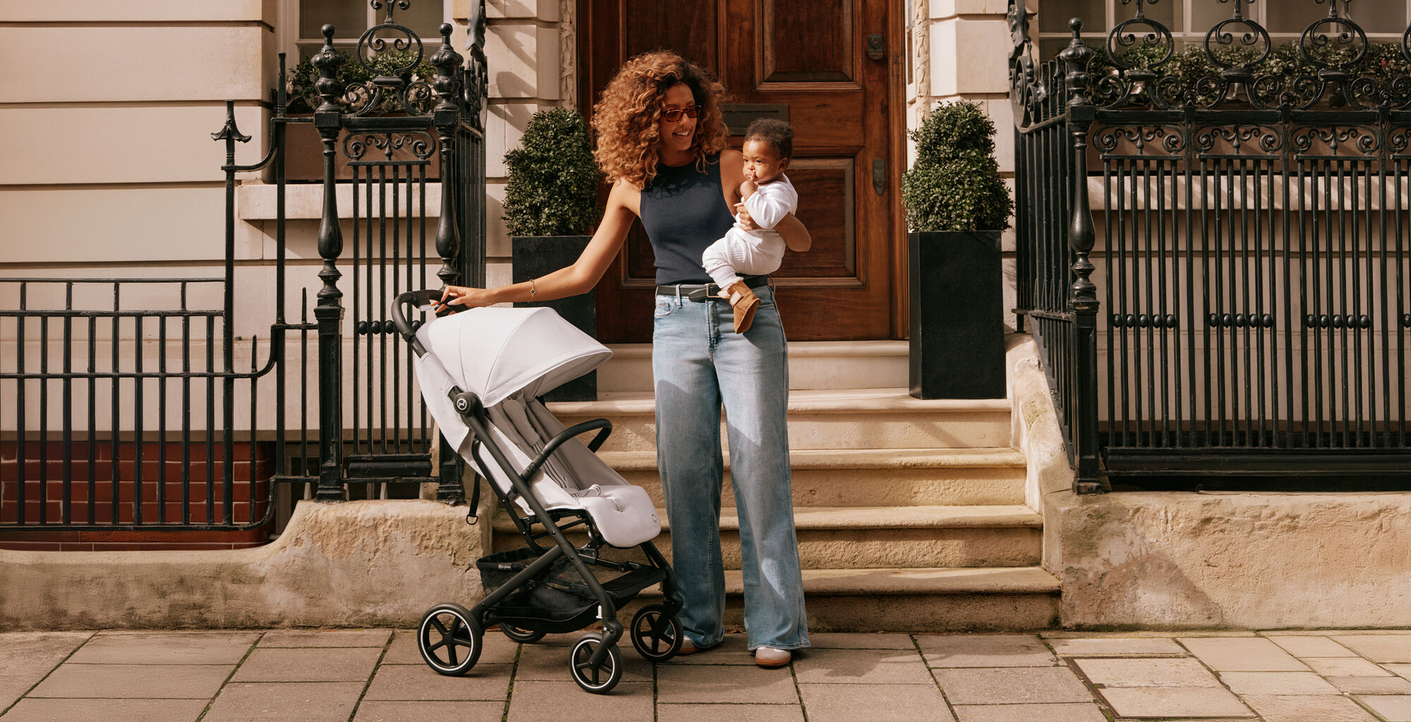 A woman stands with a baby in her arms next to a Beezy lightweight compact stroller in front of an elegant front door.