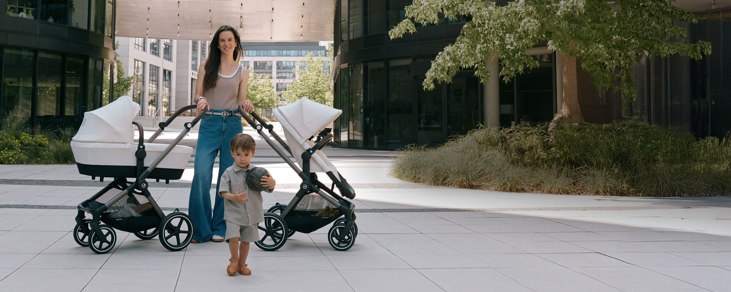 Brunh&aring;rig mamma med tv&aring; CYBEX EOS Lux barnvagnar utomhus i en somrig stadsmilj&ouml; &ndash; en barnvagn med s&auml;tesstol, en med liggdel i benvitt tyg, liten pojke med boll i f&ouml;rgrunden.