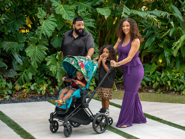 A family walking with a pram through a garden surrounded by large green leaves. The pram has a colourful canopy.