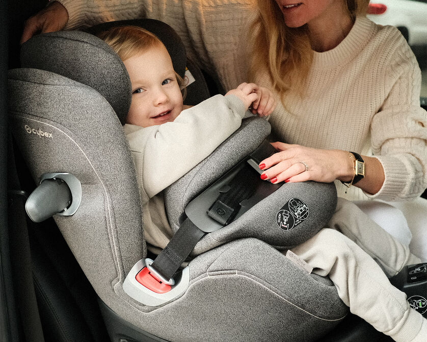 A child smiling while sitting in a Cybex car seat, as a woman in a cream jumper adjusts the seat.