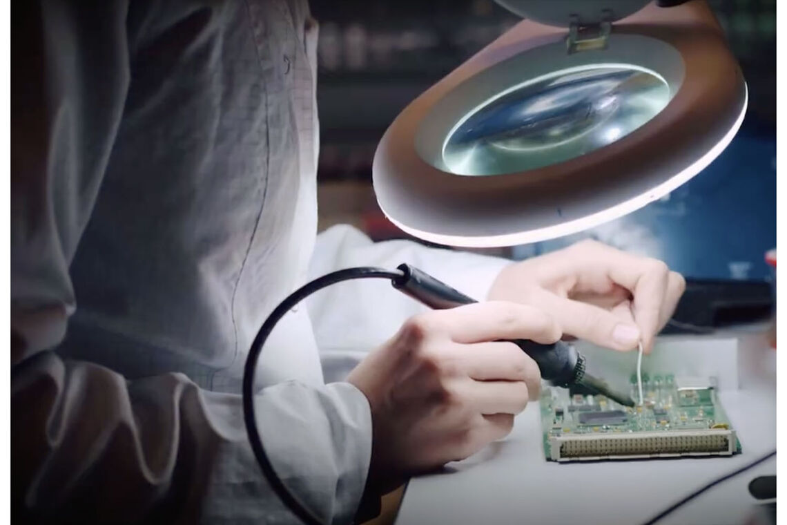 A close-up of a fair-skinned technician wearing a white lab coat sitting at a table with a magnifying glass and soldering a small green electronic circuit.