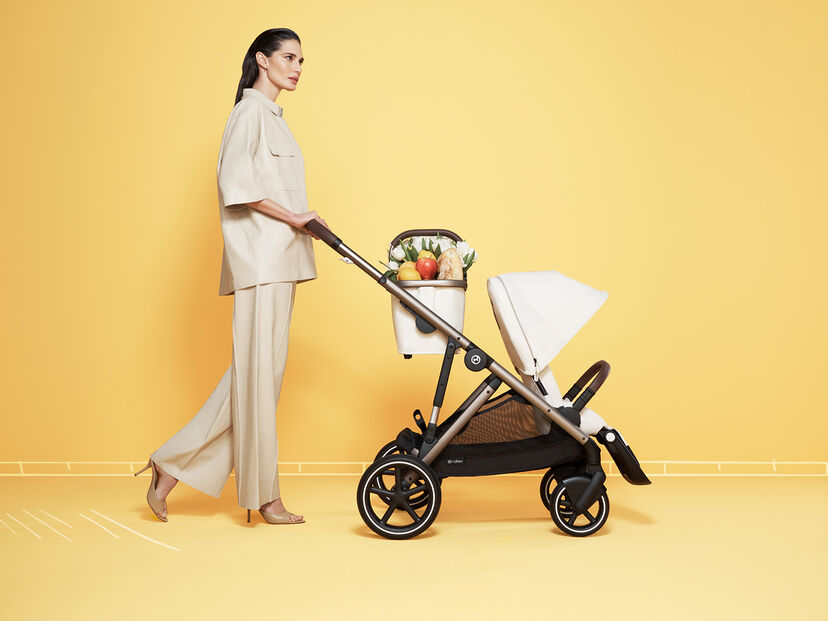 Woman in a beige outfit pushing a stroller with a grocery bag full of food against a yellow background.