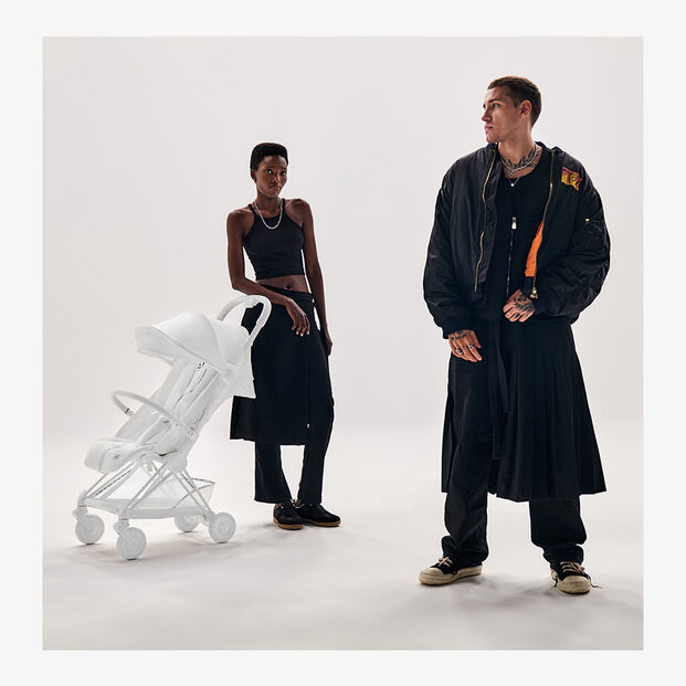 Two people in stylish black clothing stand next to a white pram in a minimalist studio.