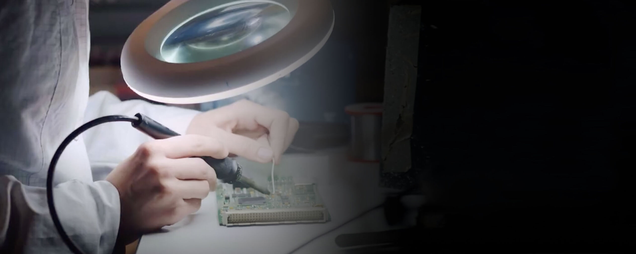 A technician soldering a circuit board under a magnifying lamp.