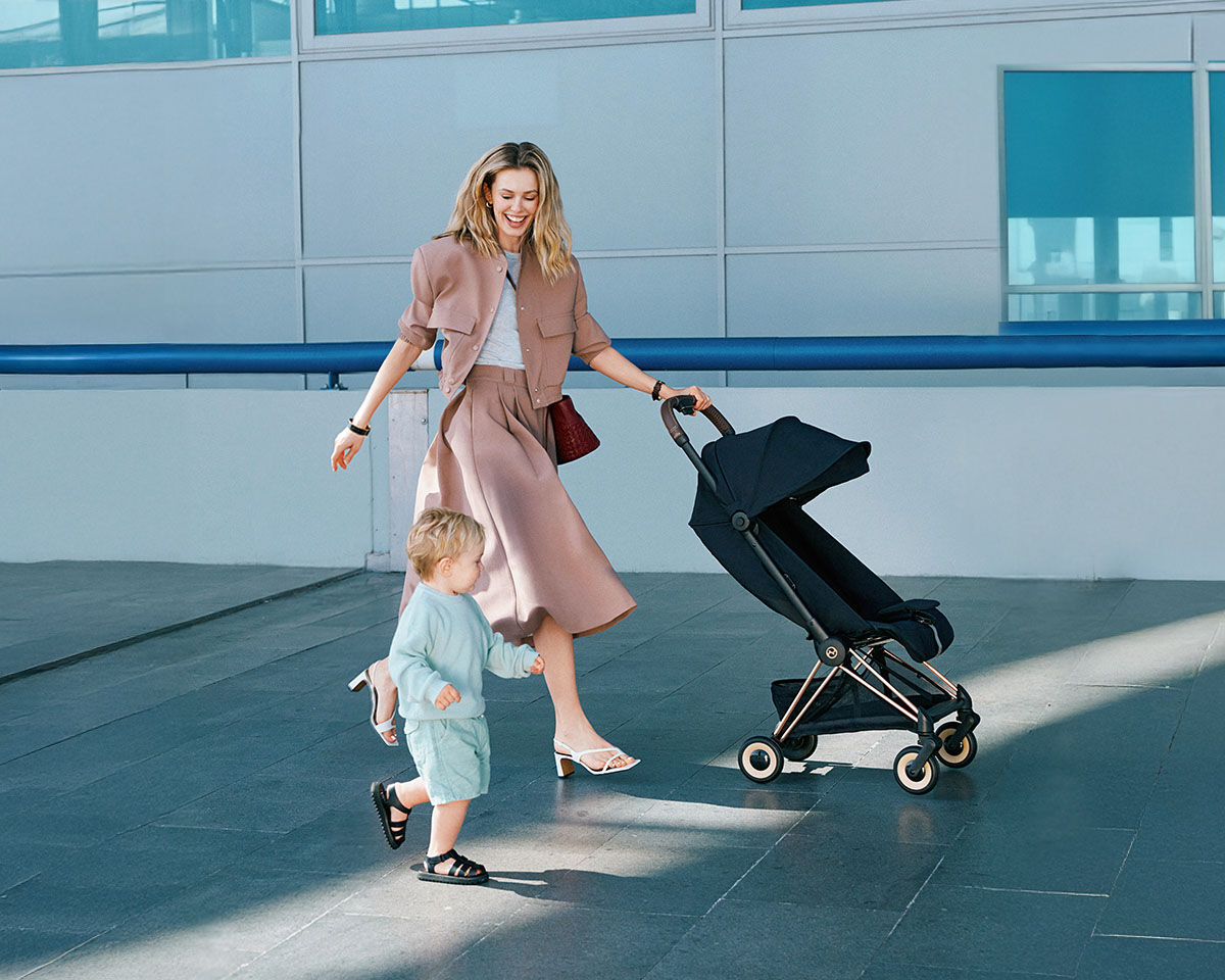 A woman in stylish clothing pushes a stroller while walking beside a small child in a blue outfit.