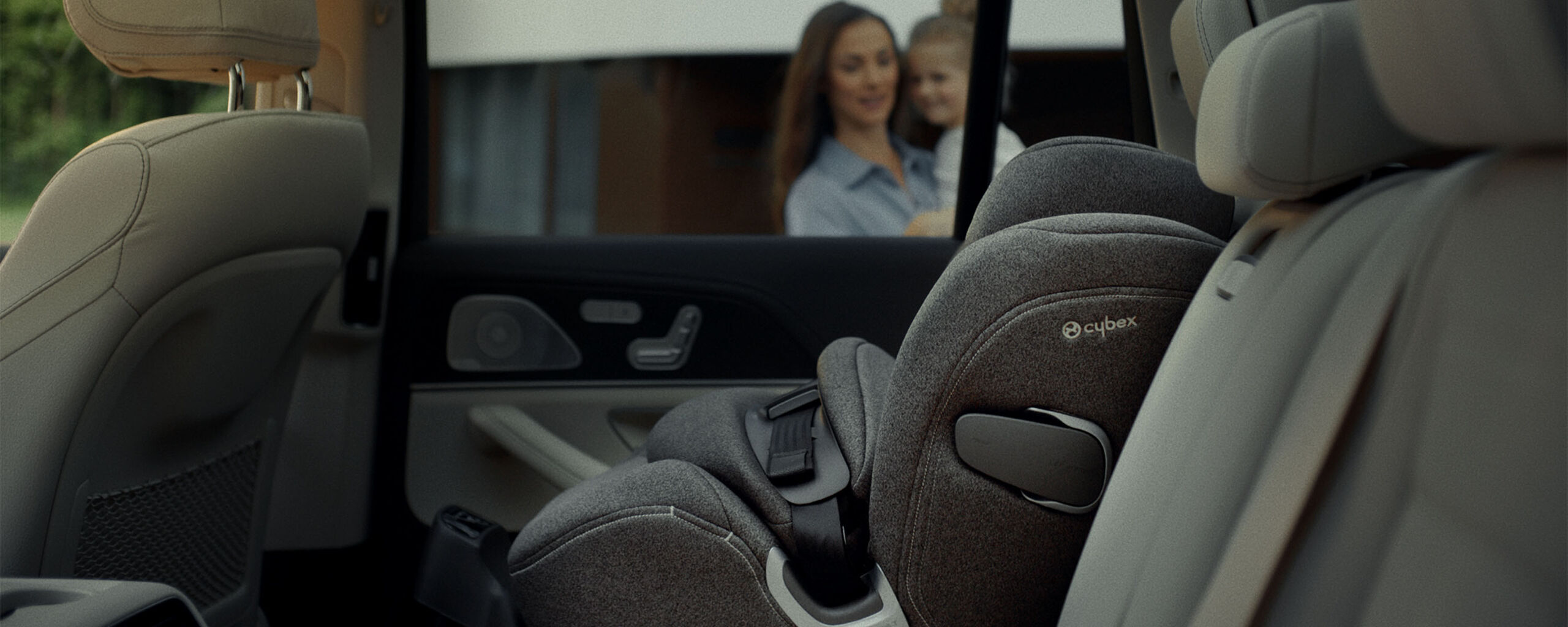 A Cybex Anoris T car seat is installed on the back seat of a car, with a woman holding a child in the background.