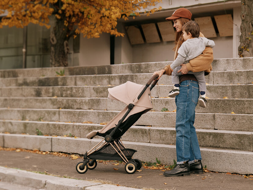 A woman stands holding a toddler next to a stroller on a street with autumn leaves.