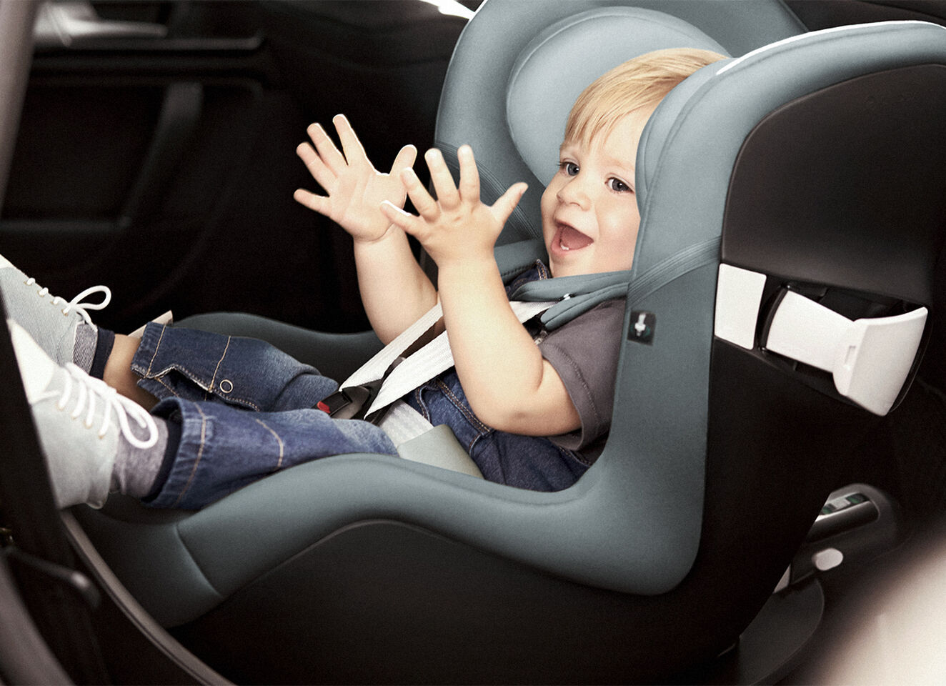 A smiling toddler sits in a car seat, joyfully reaching out with its hands.