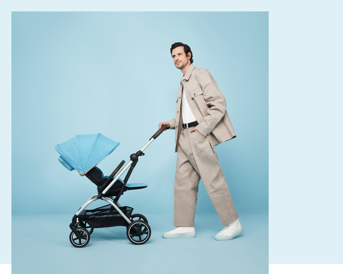 A man in a beige suit pushes a blue Eezy S Twist +2 pram against a light blue background.