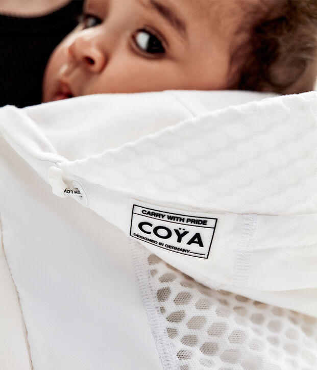A baby looks over an adult's shoulder, wrapped in a white fabric with the label "CO&Yuml;A, Designed in Germany".