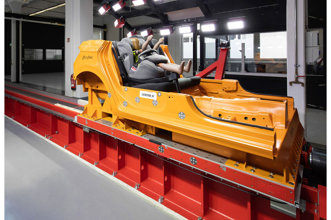 An orange car segment with a grey Cybex car seat and dummy doll mounted on it, set on rails in a crash test lab.