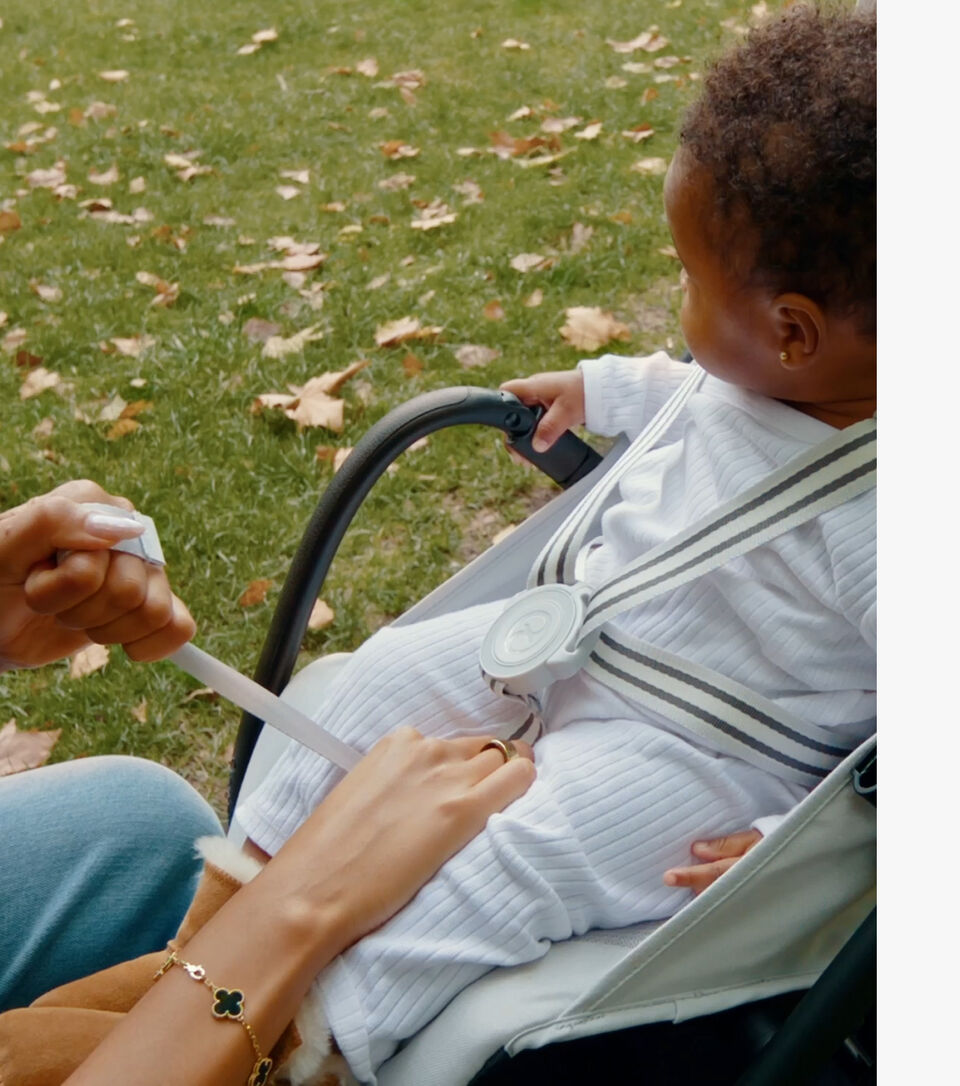 A woman with curly hair holds a baby in a park, next to her is the Beezy lightweight compact stroller.