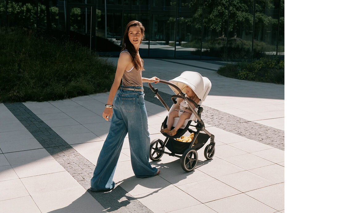 En brunh&aring;rig kvinna skjuter CYBEX EOS LUX barnvagn i Seashell Beige d&auml;r ett sm&aring;barn sitter. Bilden ing&aring;r i en kampanjbild tagen utomhus.