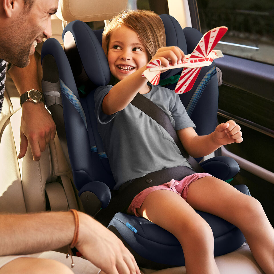 A child happily sits in a Solution S2 car seat, playing with a toy airplane, while an adult smiles at them.