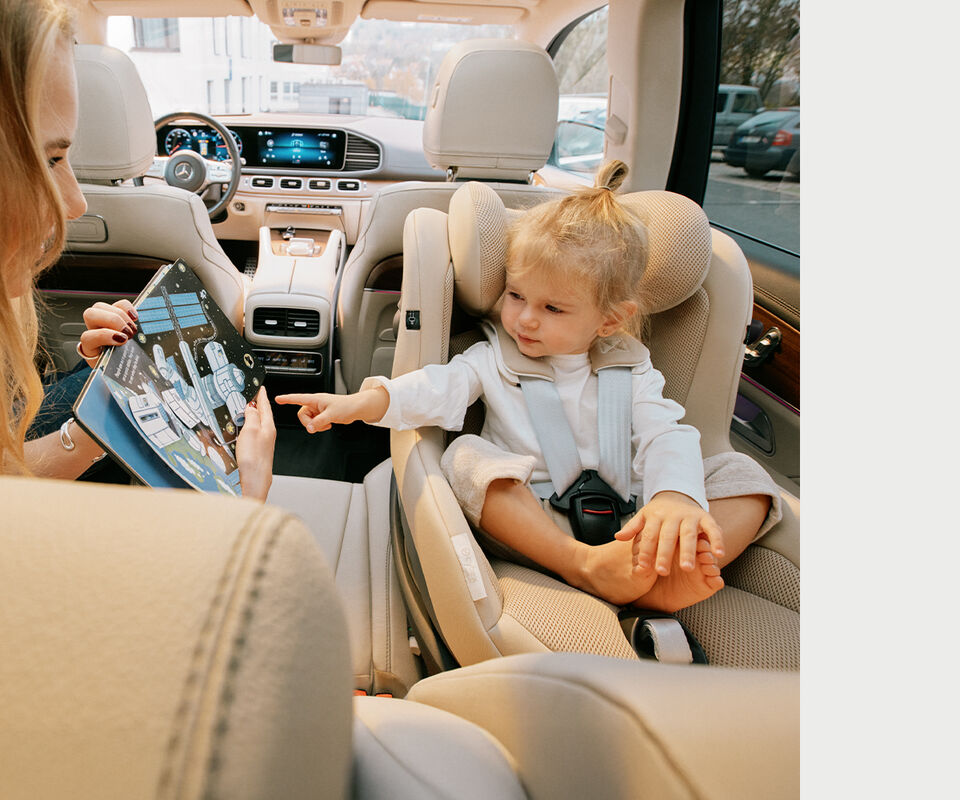 Child pointing at a picture book the mother is holding up, the child sitting in the back of a car in a Sirona G i-Size car seat.