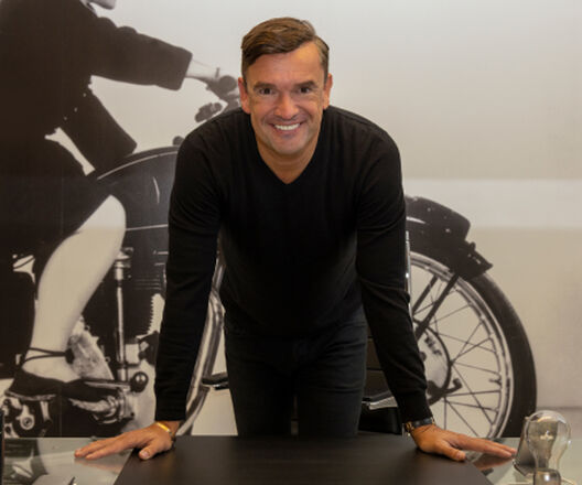 A man in a black jumper smiles as he leans over a desk, with a large black-and-white photograph of a vintage motorcycle behind him.
