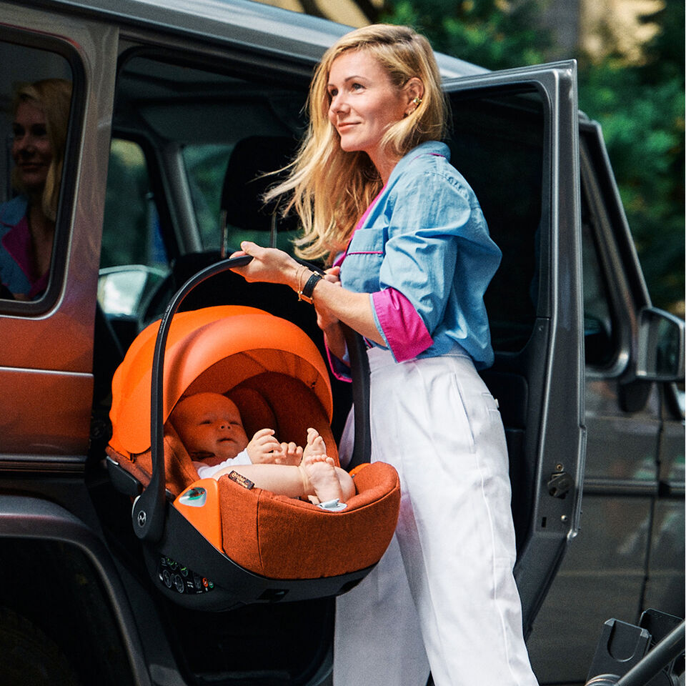 Woman with blonde hair lifting an orange car seat with a baby out of a car.